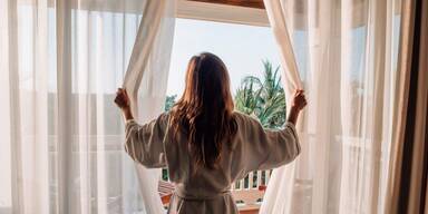 Frau steht vor Fenster und &ouml;ffnet Gardine