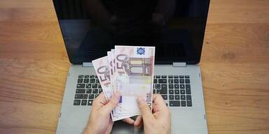 Aufgeklappter Laptop, davor M&auml;nnerh&auml;nde die vier F&uuml;nfzigeuroscheine halten.