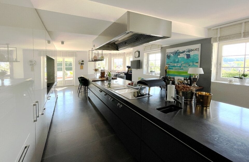 Hochmodern und geräumig: Die Küche ist top ausgestattet und wirkt edel. Zu den weißen Fronten gibt es mit der dunklen Kochinsel einen starken Kontrast, einziger Farbklecks ist das Gemälde an der Wand.